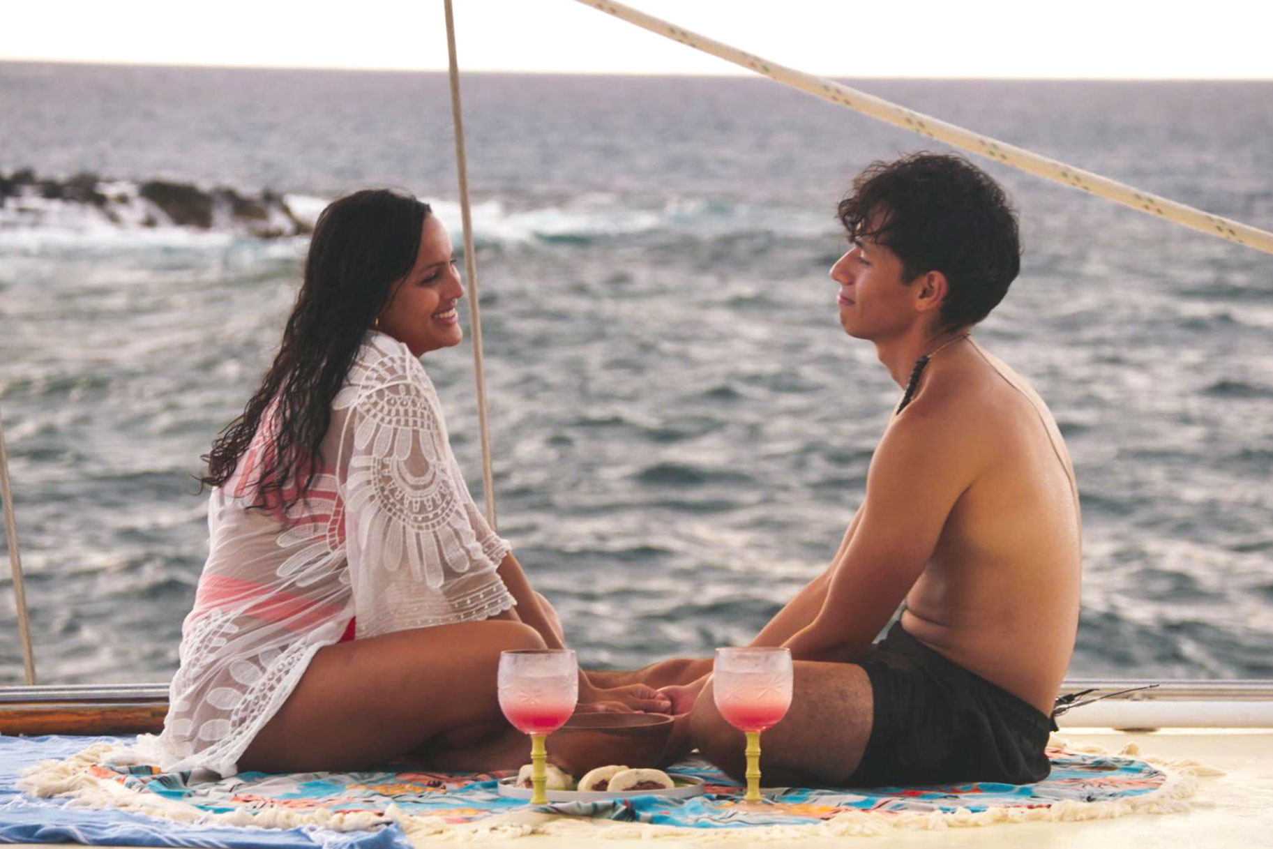 Edgar and Marissa sit facing each other in front of the ocean, holding hands