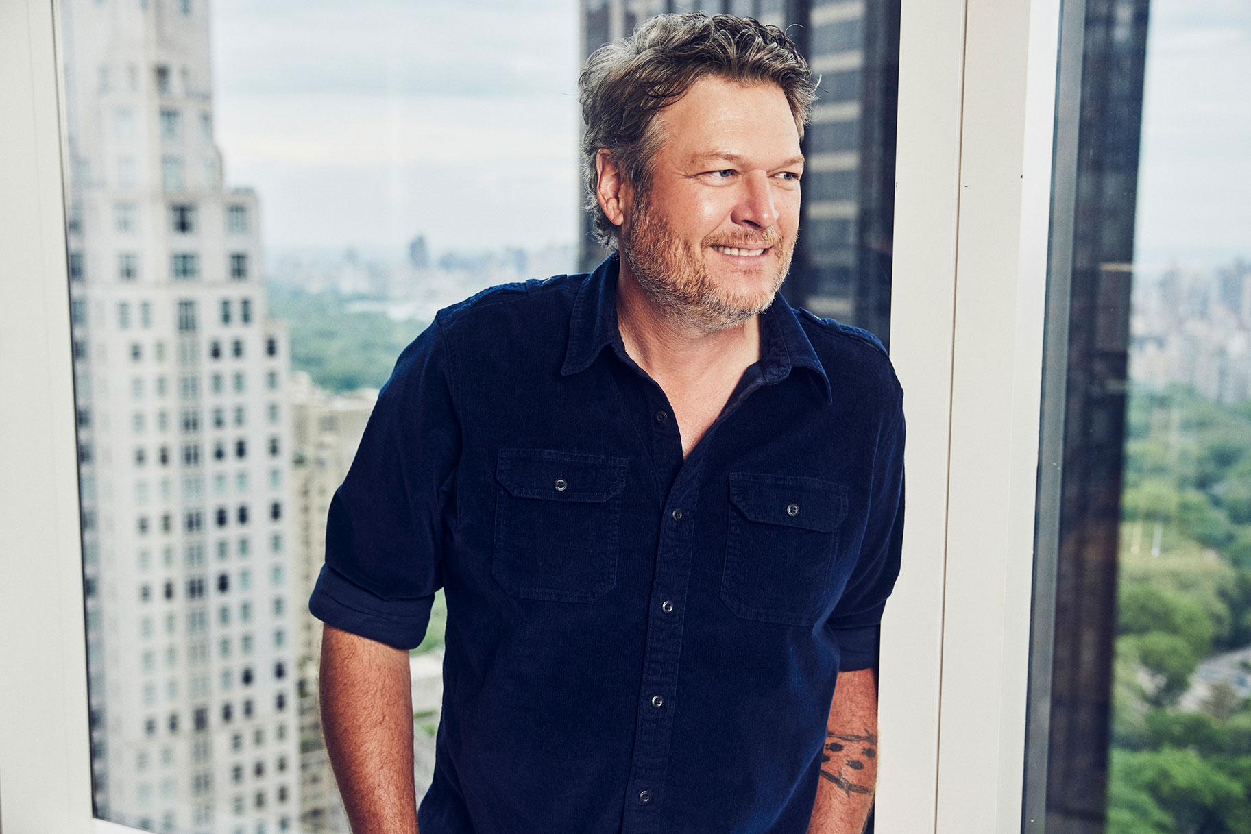 Blake Shelton standing in front of large windows overlooking a big city