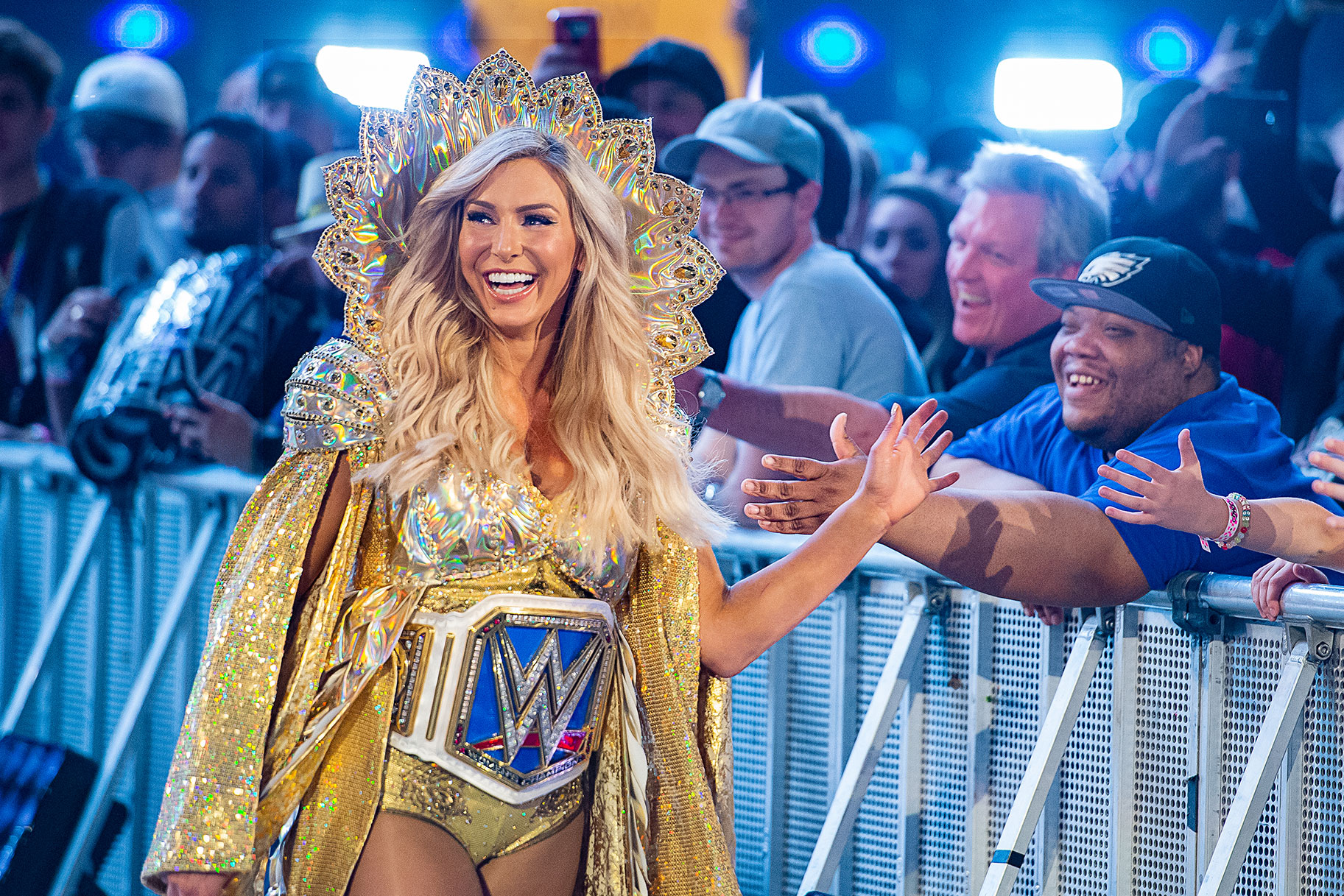 Charlotte Flair walking into the WWE ring wearing an extravagant gold jacket and headpiece. She is smiling