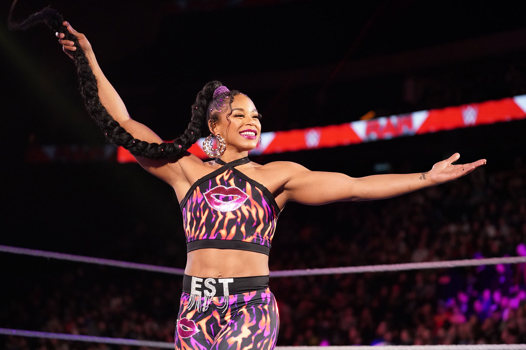 Bianca Belair holding her long braid in her right hand with both arms outstretched. She is smiling.