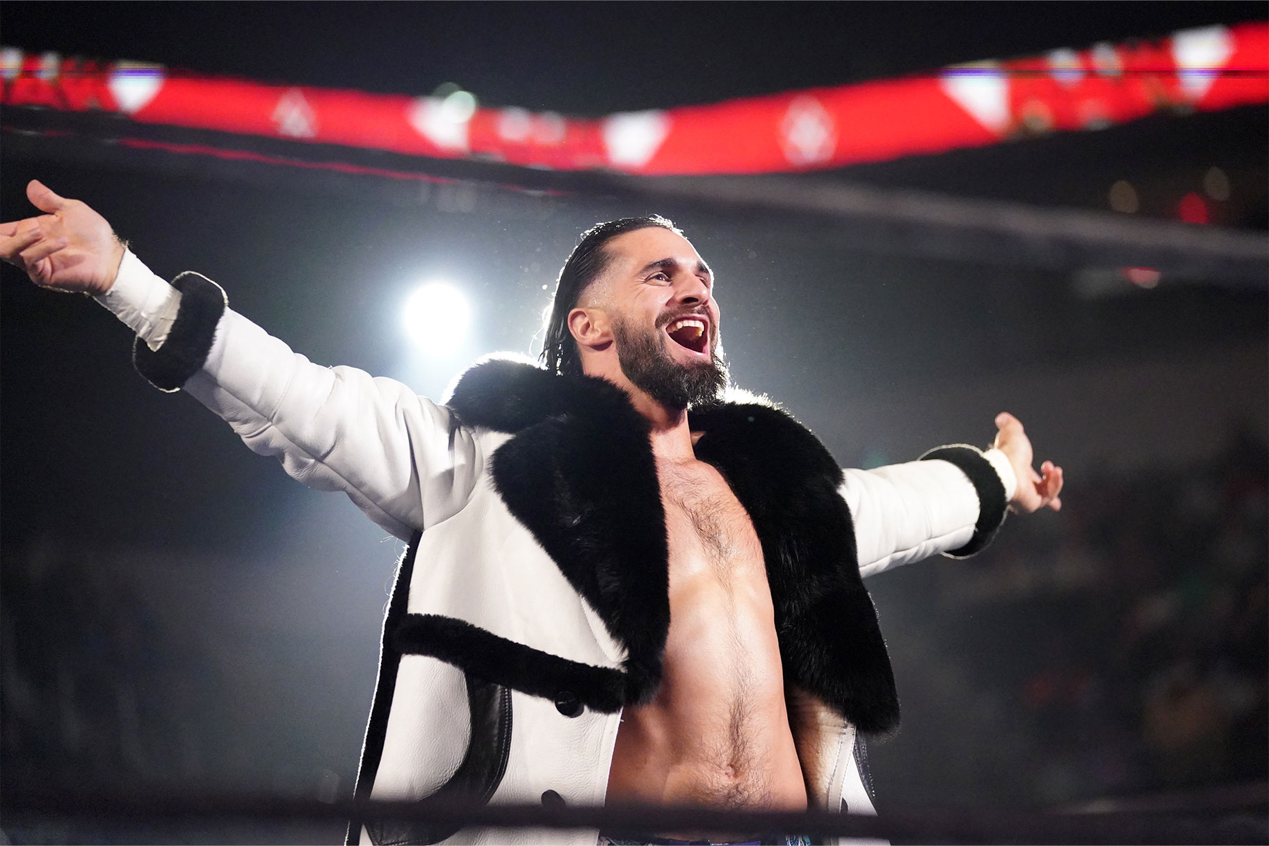 Seth Rollins wearing a white coat in the ring, smiling with his arms outstretched
