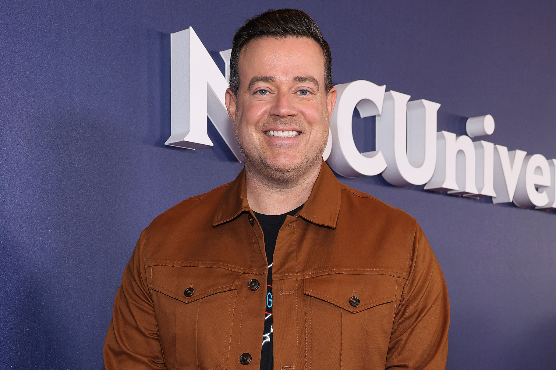 Carson Daly smiling as he stands on an NBC red carpet