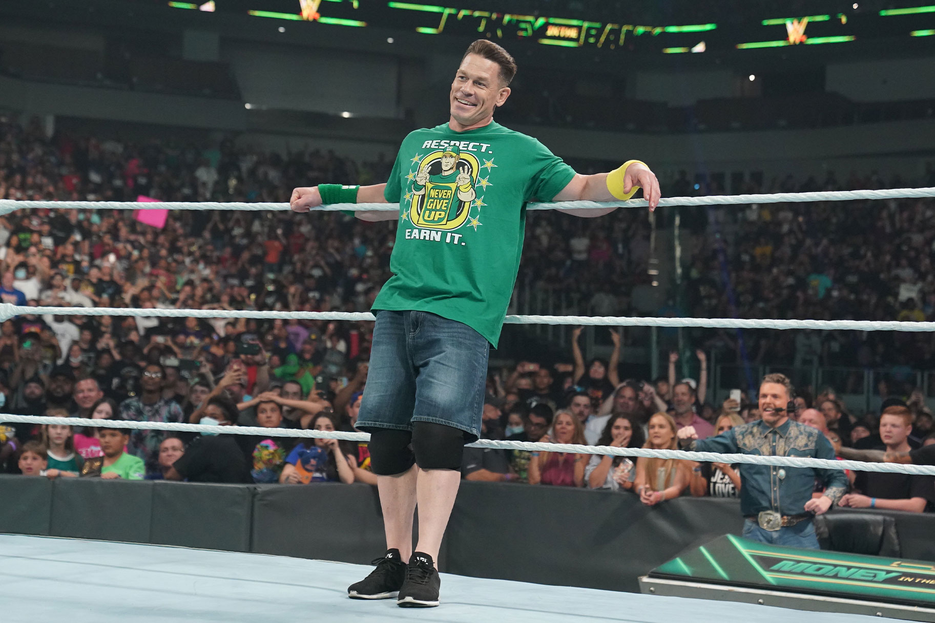 John Cena leaning against the top rope, wearing a green shirt and denim shorts