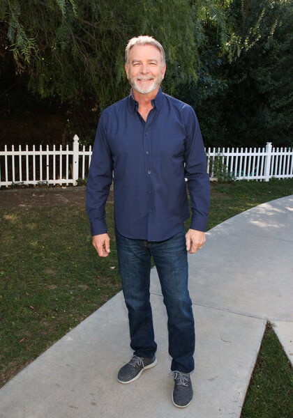 Bill Engvall full length wearing a blue button down shirt and jeans while in a yard setting.