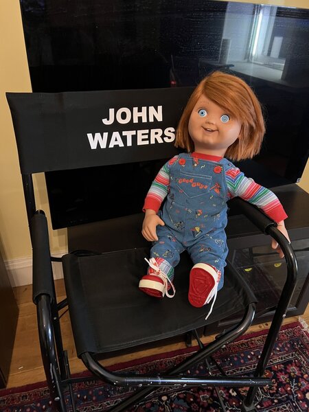 Chucky sits on a chair marked "John Waters".