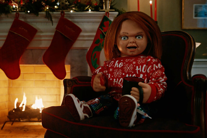 Chucky wearing a red Christmas sweater in front of a fireplace