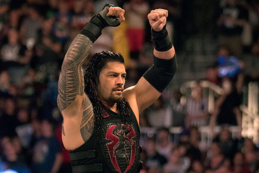 Roman Reigns victoriously puts his fists in the air while standing on the top rope.