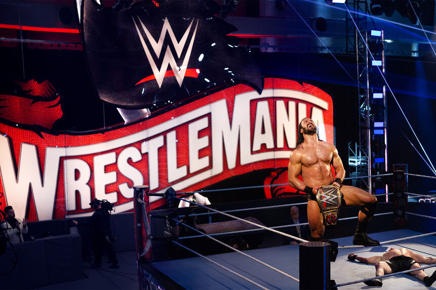 Drew McIntyre on. the top of the rope holding a belt while a large Wrestlemania logo can be seen behind him